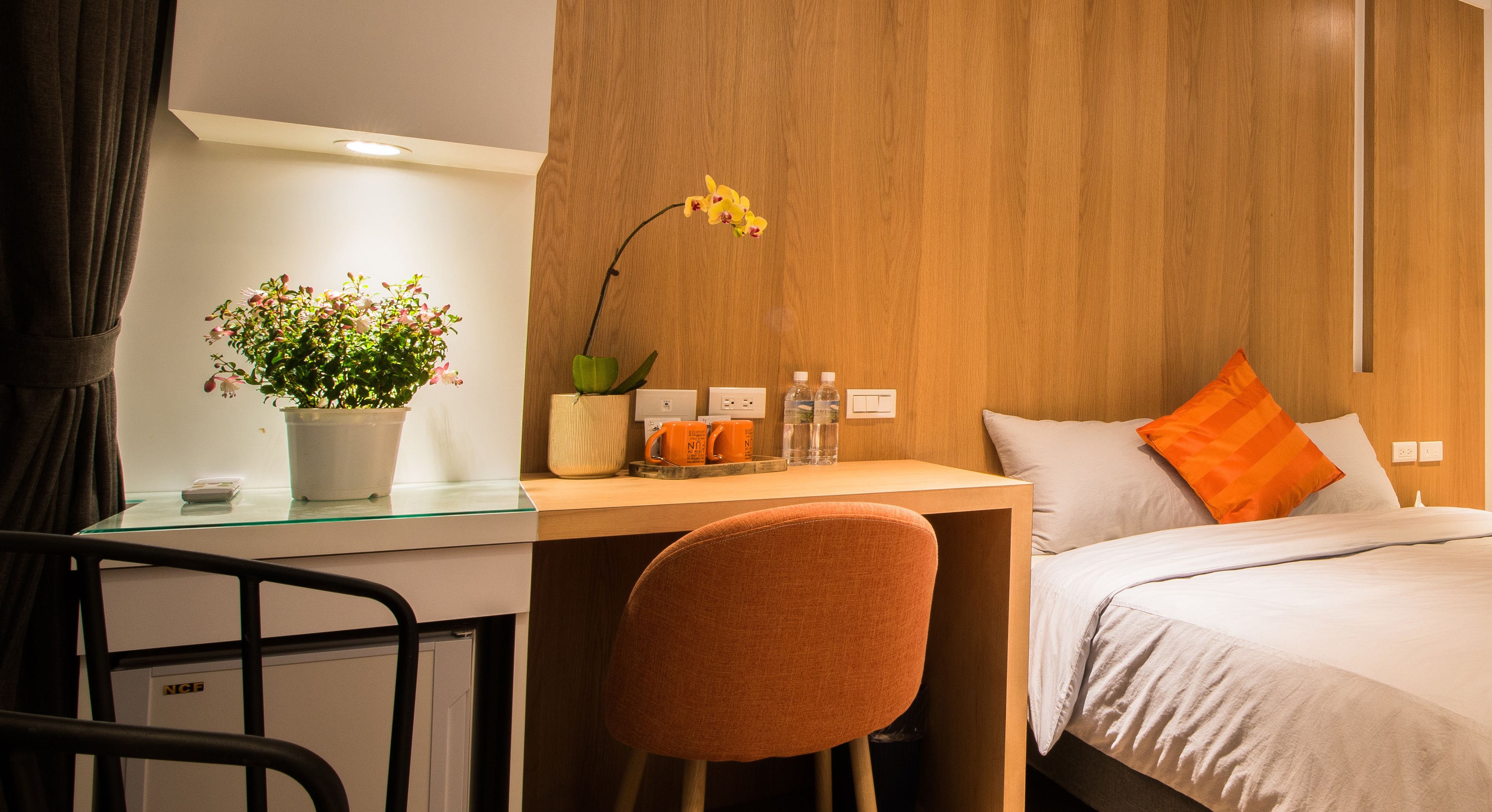 Hotellihuone, jossa on sänky, työpöytä tuolilla, ruukkukasvi, lamppu sekä yöpöytä, jossa on lamppu ja lasi.