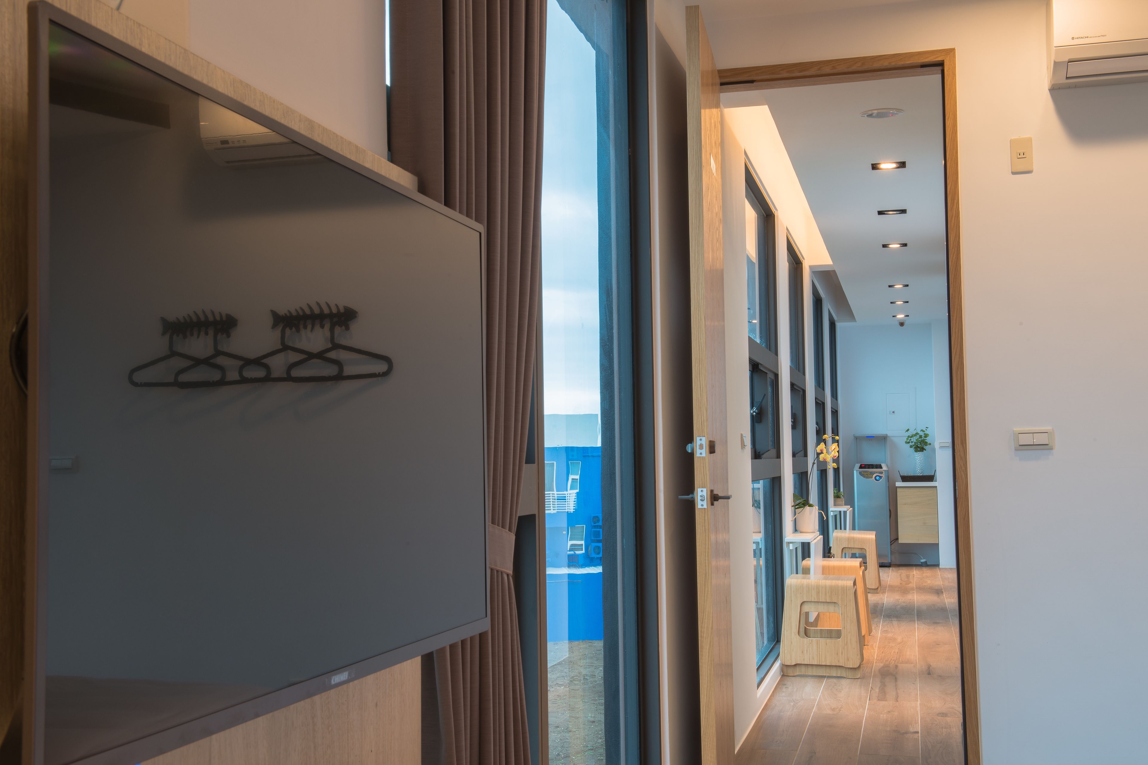 En moderne hotelgang med trægulv, en glasdør med metalgreb og udsigt ind til et værelse med seng og skrivebord.
