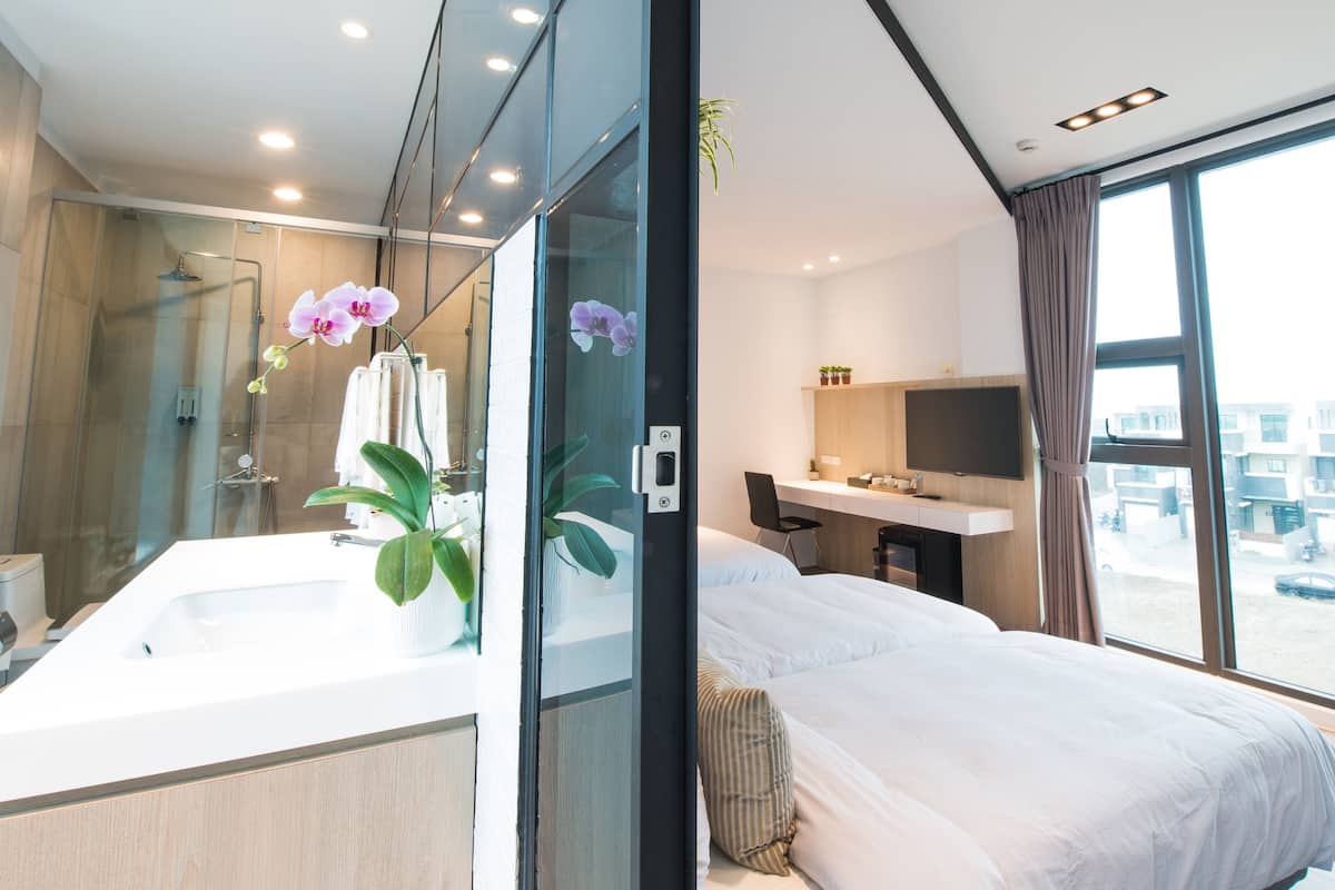 A modern hotel room with a large bed, a separate bathroom with a shower, and a view of the city through large windows.