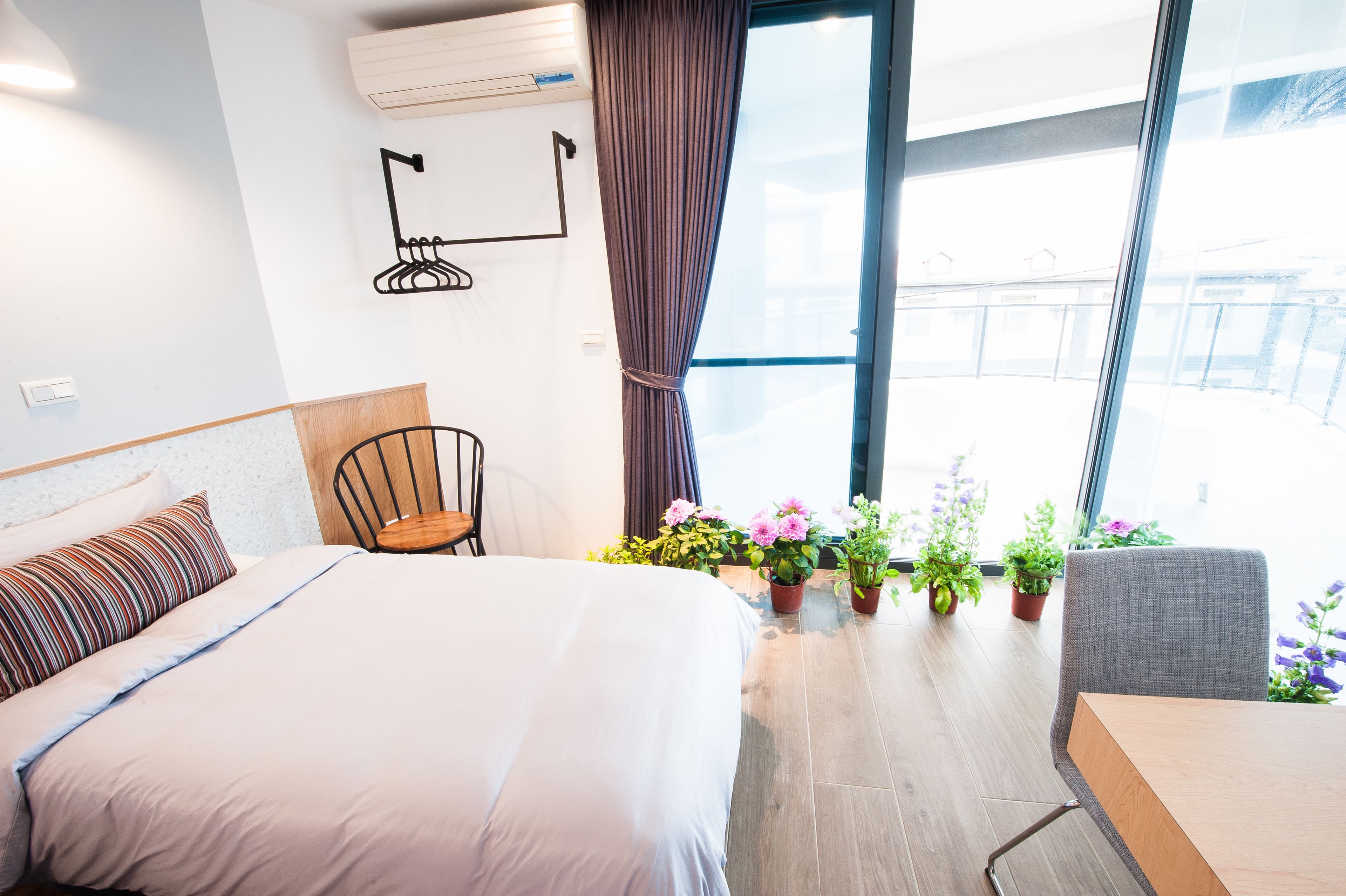A modern hotel room with a large bed, a desk, and a chair. There are potted plants and a view of the outside through large windows.