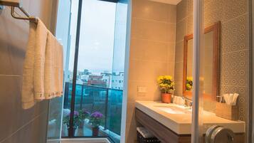 A modern bathroom with a glass shower door, a large window offering a city view, and a vanity with a mirror and sink.