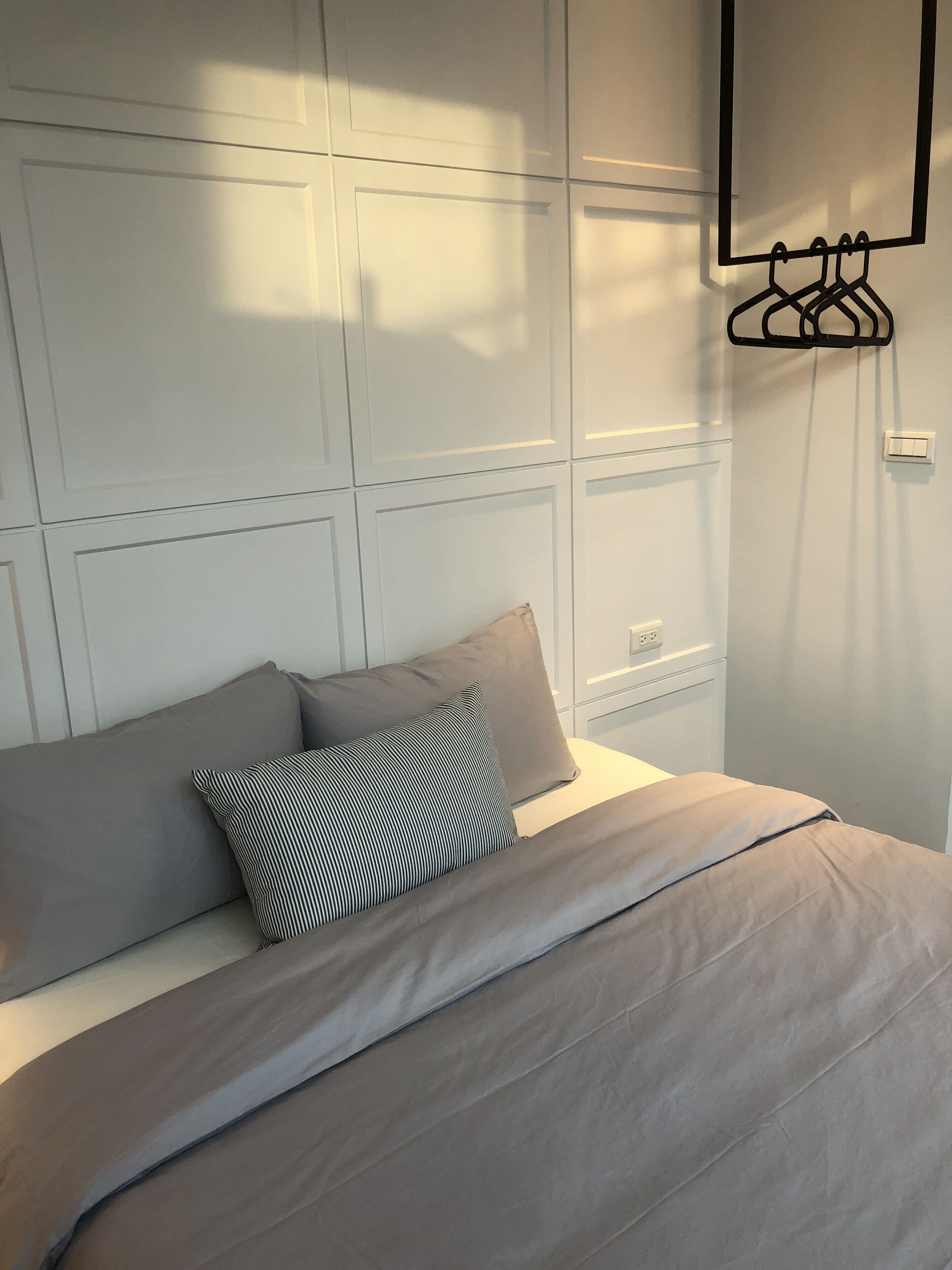 A neatly made bed with a grey duvet and pillows, a wall-mounted light switch, and a black hanger rack.