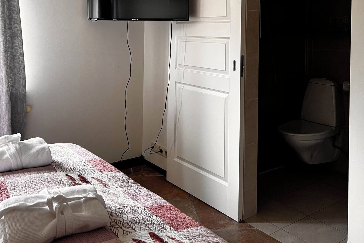 A bedroom with a bed, a flat-screen TV mounted on the wall, a door leading to a bathroom, and a curtain on the window.