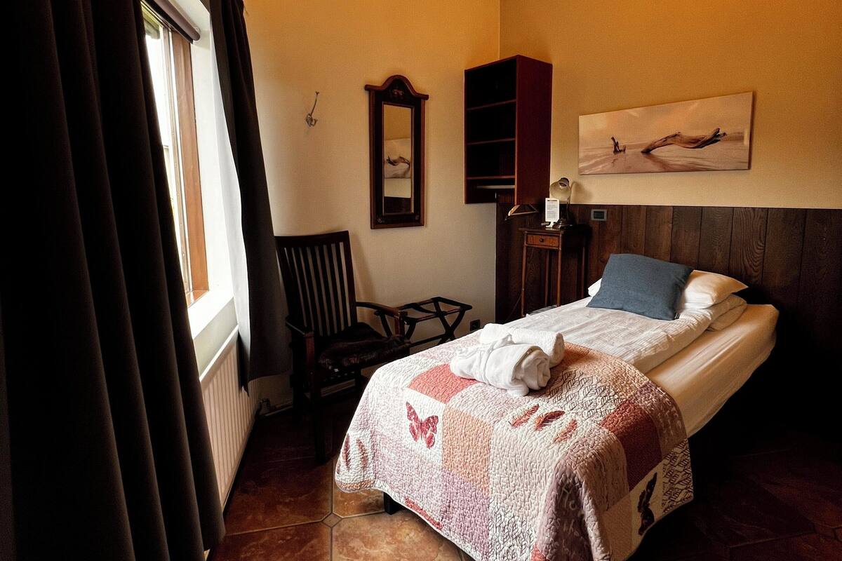 A bedroom with a bed, a chair, a wooden cabinet, a painting, and a chandelier.