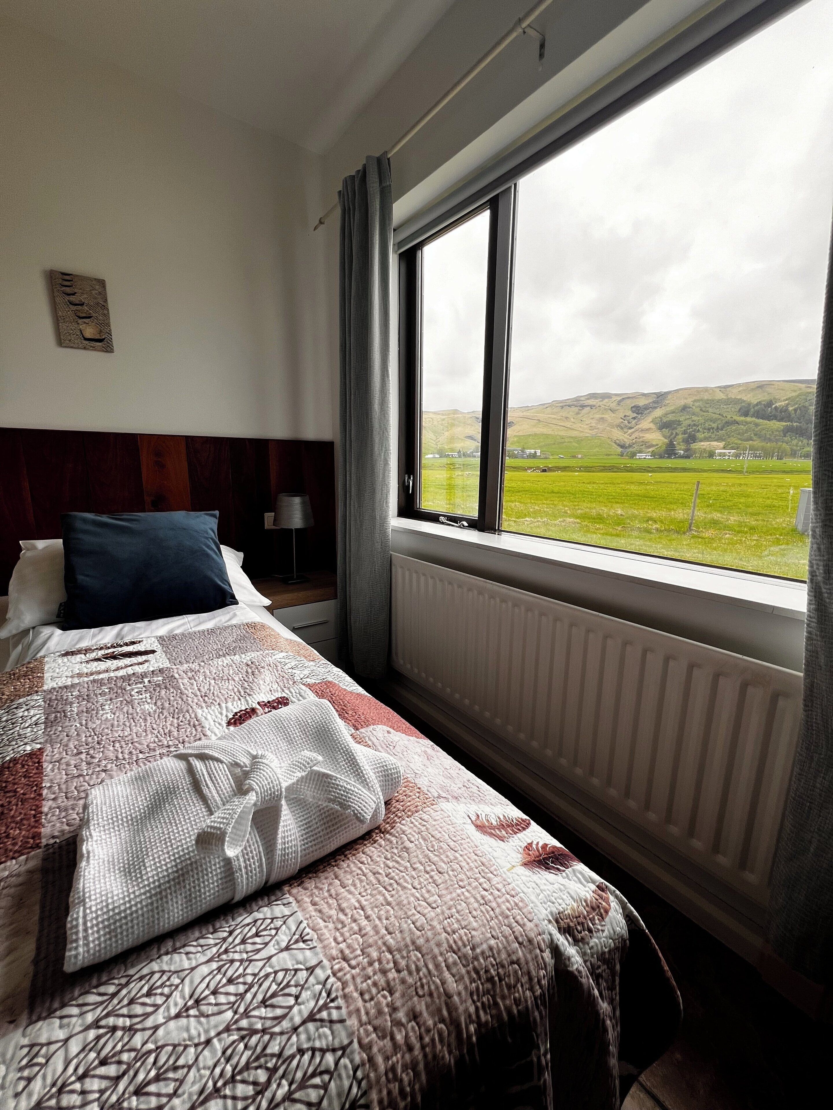 A bedroom with a large window, a bed with a patterned quilt, a radiator, and a view of a green landscape.