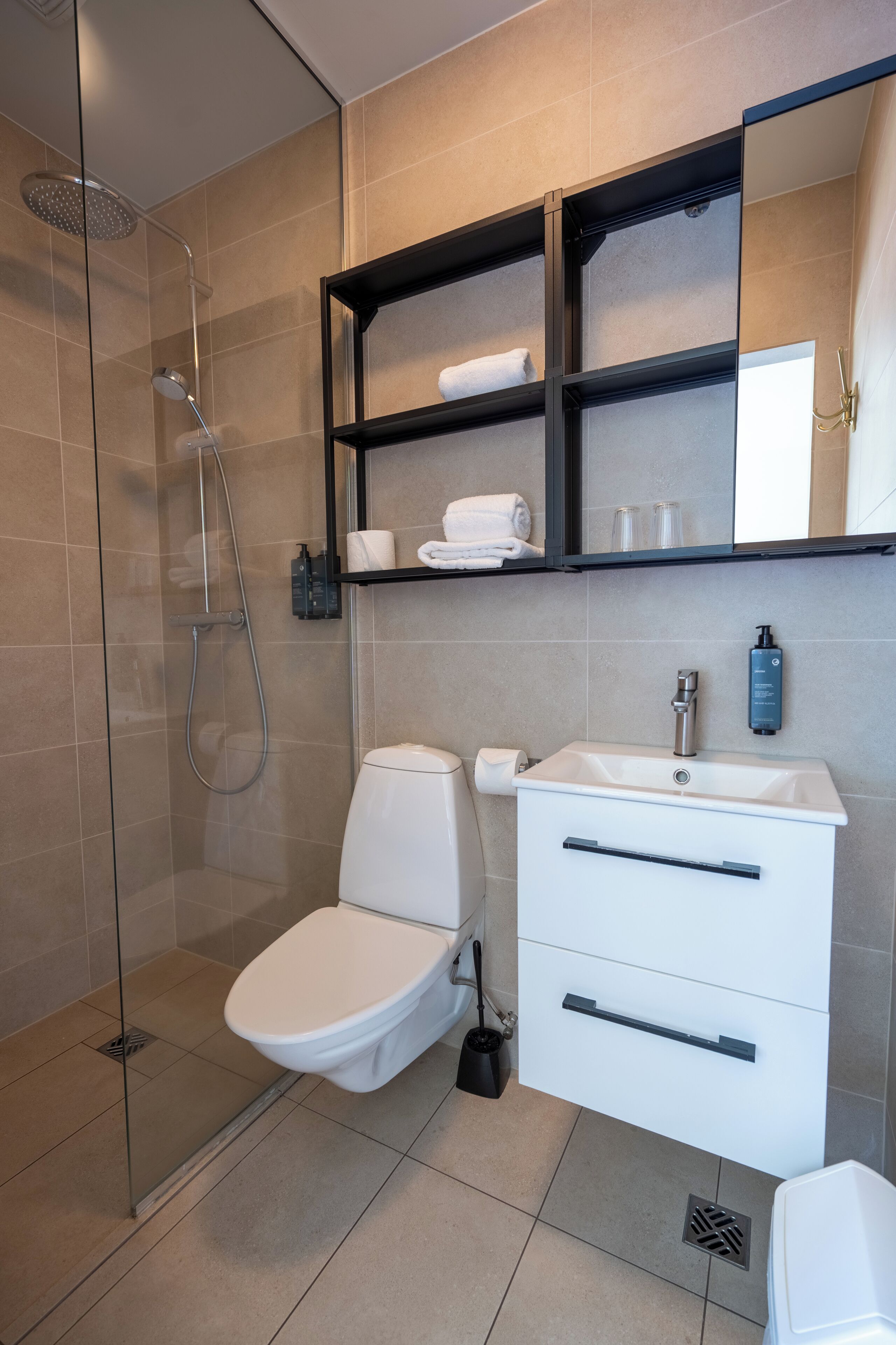 Un baño moderno con cabina de ducha de vidrio, inodoro blanco, lavamanos blanco con espejo y estante con toallas y artículos de aseo.