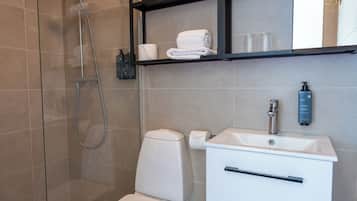 Un baño moderno con cabina de ducha de vidrio, inodoro blanco, lavamanos blanco con espejo y estante con toallas y artículos de aseo.