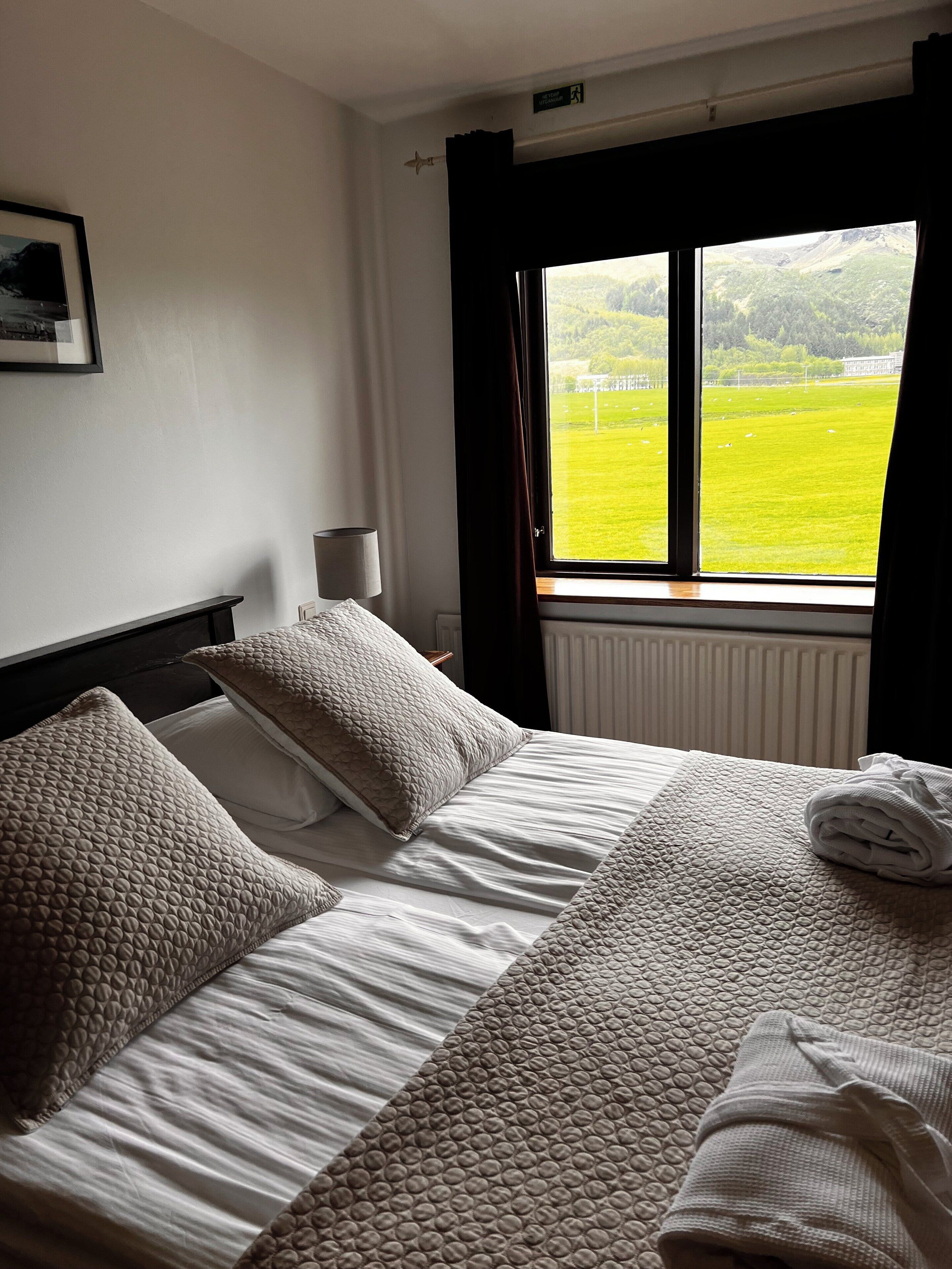 A hotel room with a bed, pillows, a lamp, and a window with a view of a green field.
