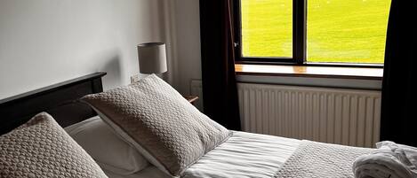 A hotel room with a bed, pillows, a lamp, and a window with a view of a green field.
