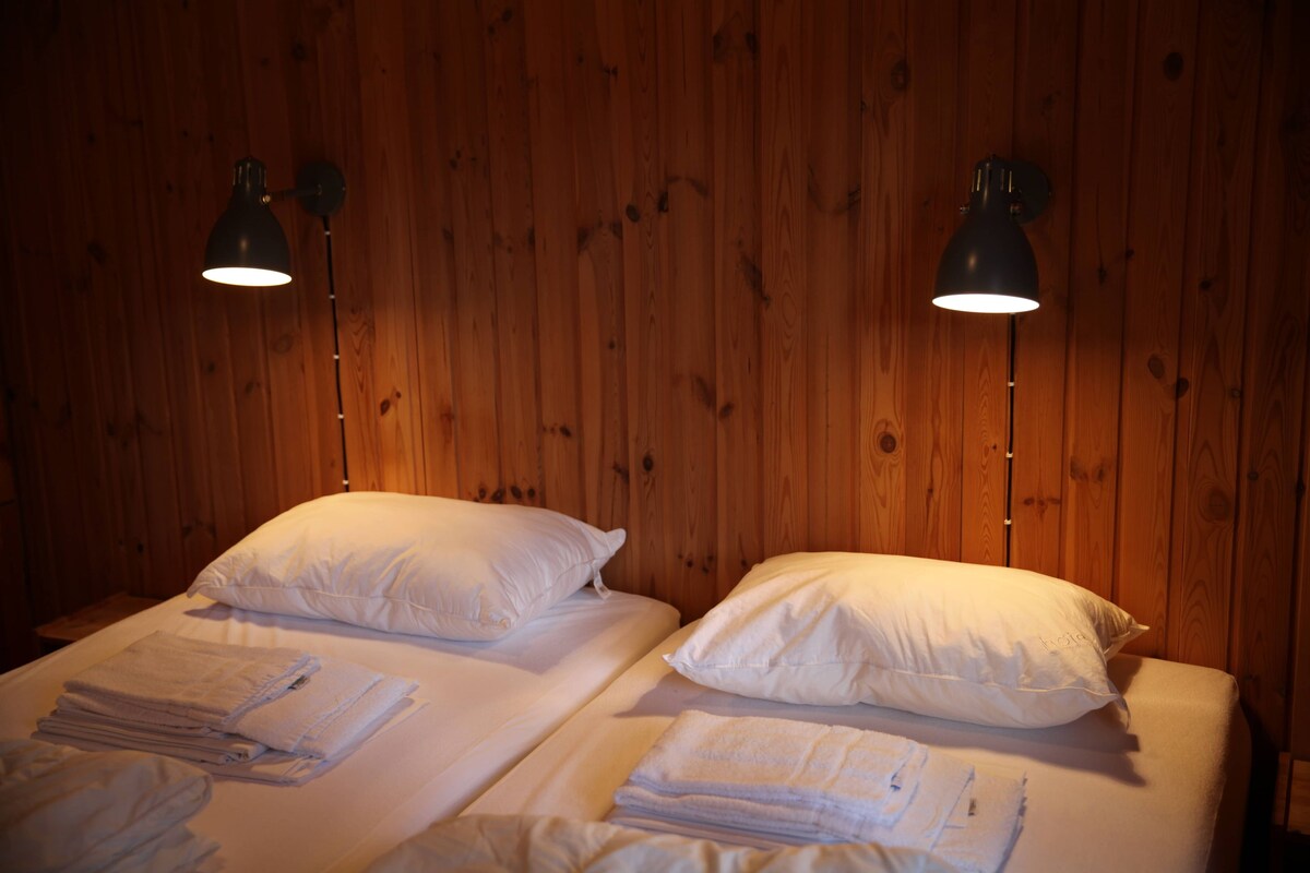 Dos camas con sábanas y almohadas blancas, un panel de madera y dos lámparas de pared.