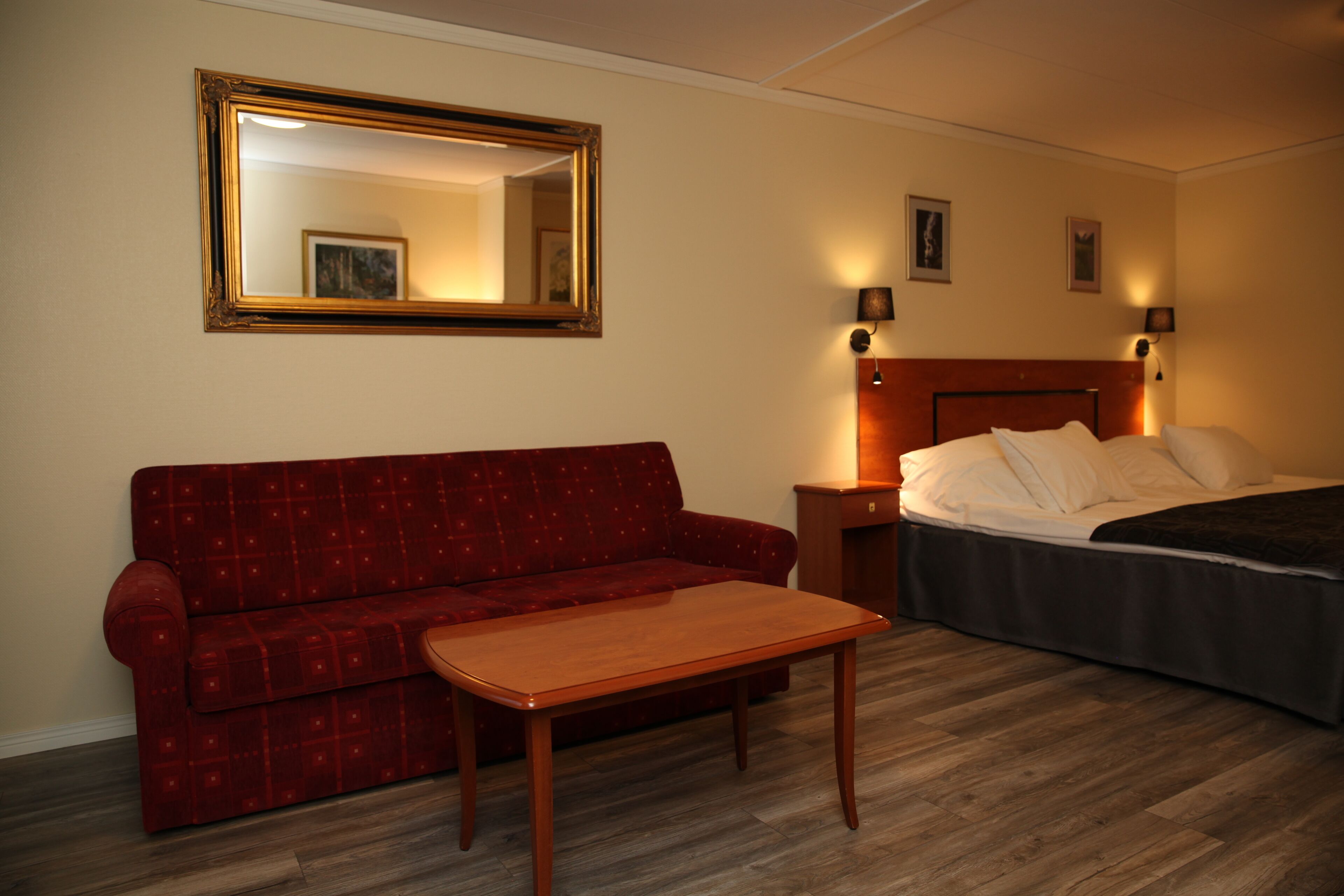 A hotel room with a red sofa, a wooden table, a bed with white bedding, a nightstand, and a wall-mounted mirror.
