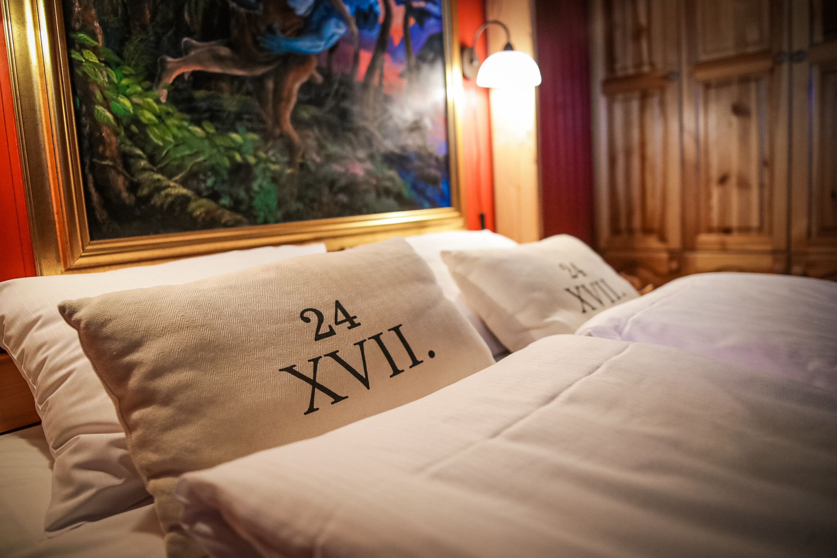 Una cama con almohadas etiquetadas '24 XVII' en una habitación con cabecera de madera y un cuadro de una escena boscosa.
