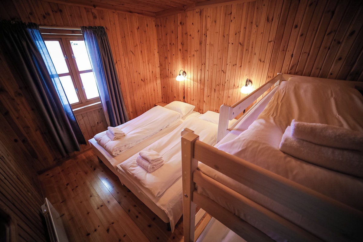 A wooden cabin room with two beds, a window with curtains, and wall-mounted lights.