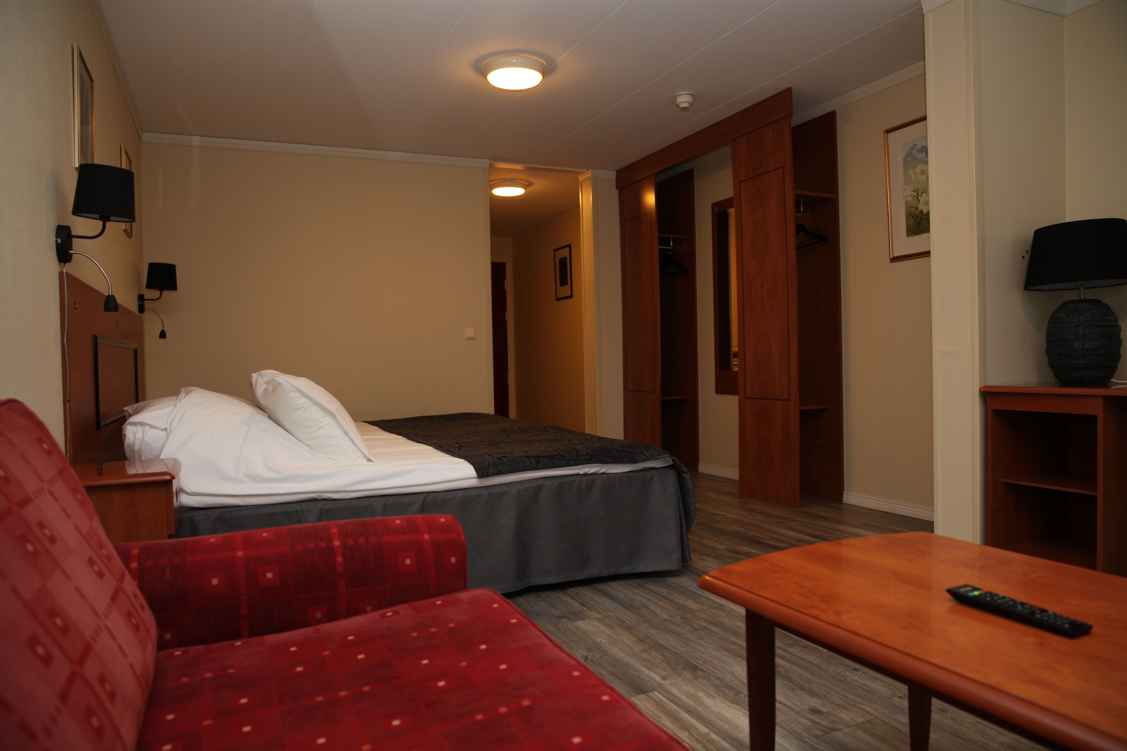A hotel room with a bed, a red sofa, a wooden table, and a remote control.