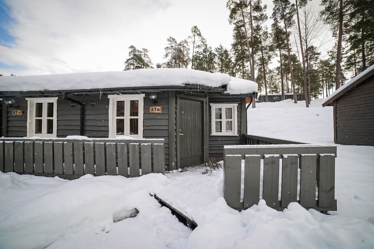 A small cabin with a snow-covered roof and a wooden fence in a snowy forest.