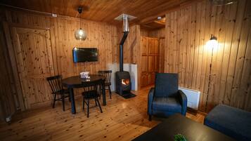 A cozy room with wooden walls, a round table with chairs, a sofa, a TV mounted on the wall, and a wood-burning stove.