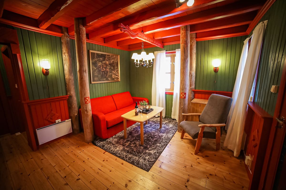 A cozy room with a red sofa, a wooden table, and a chair. There is a chandelier hanging from the ceiling and a painting on the wall.
