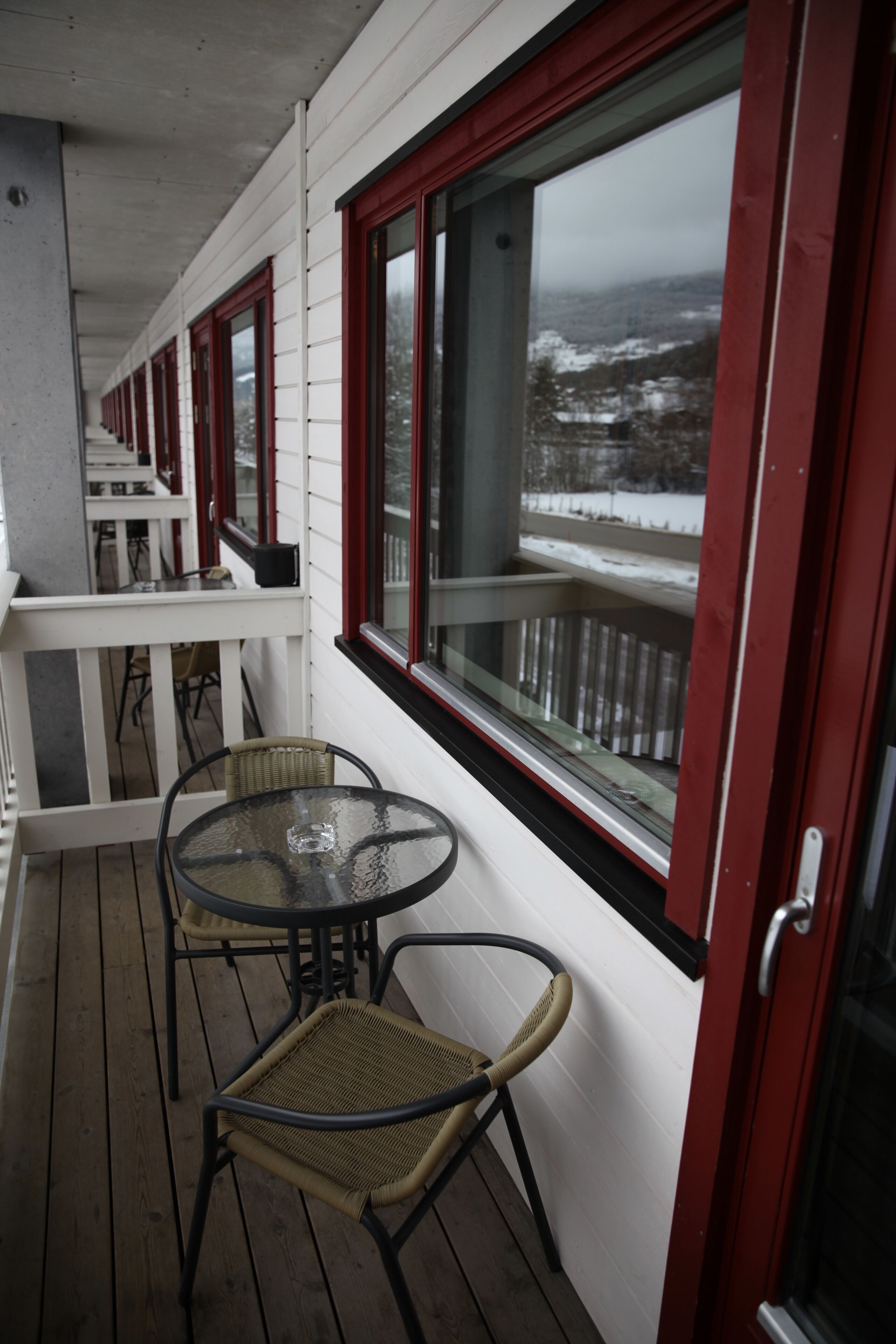 A covered outdoor area with a wooden deck, white railings, and red-framed windows.