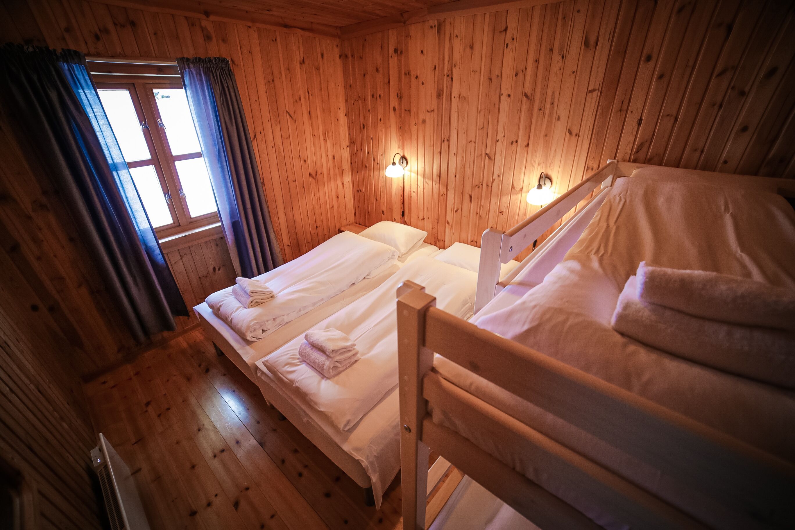 A wooden cabin room with two beds, a window with curtains, and wall-mounted lights.
