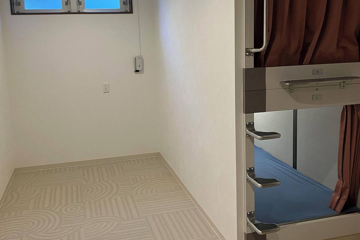 Habitación de hotel con literas, una ventana pequeña con cortinas, un enchufe y una alfombra estampada.