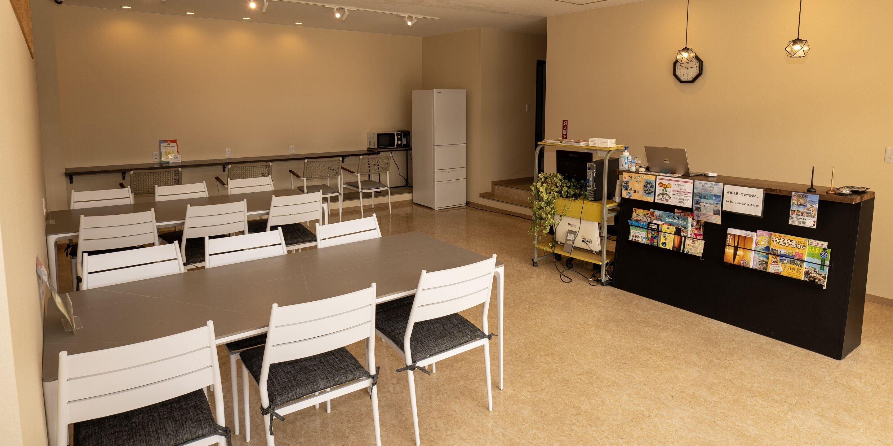 A hotel room with a dining table, chairs, a refrigerator, a microwave, a desk with a computer, and a bulletin board with various notices.