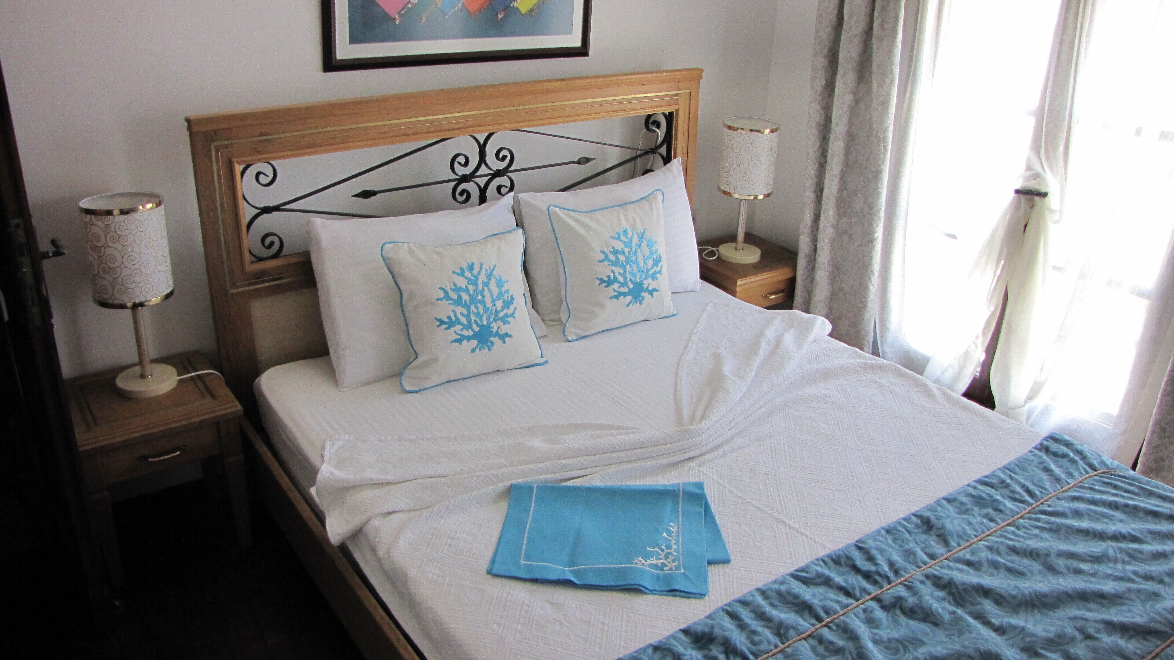 Una cama bien tendida con ropa blanca, dos almohadas decorativas, un plaid azul y dos lámparas de noche sobre mesitas de madera.