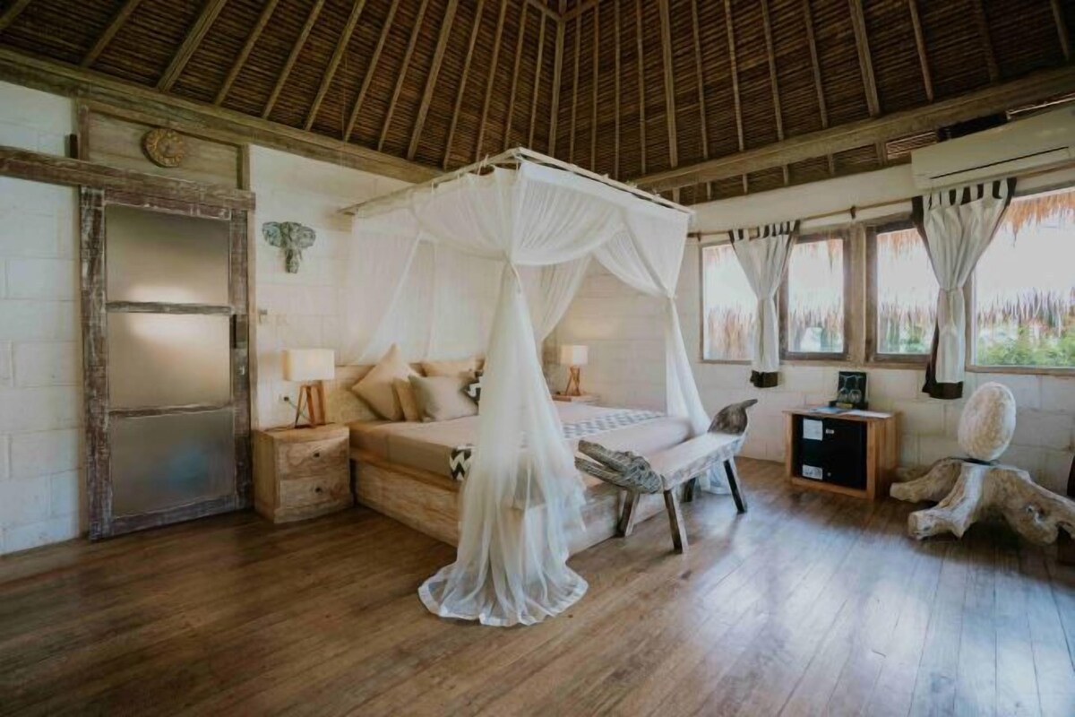 A bedroom with a canopy bed, wooden flooring, a rustic wall, and a window with curtains.