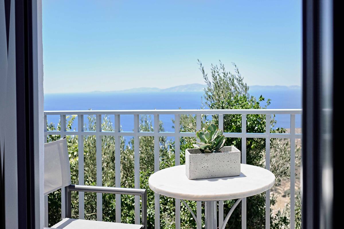 Een balkon met een witte reling, een ronde tafel met een plant in pot, en uitzicht op de zee en verre bergen.