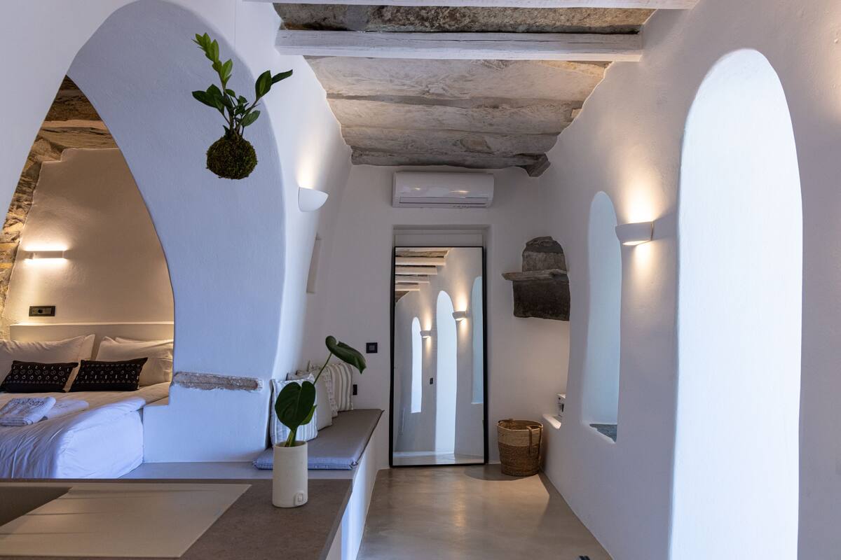 Een moderne, minimalistische hotelkamer met een bed, zithoek en een gang naar een andere kamer.