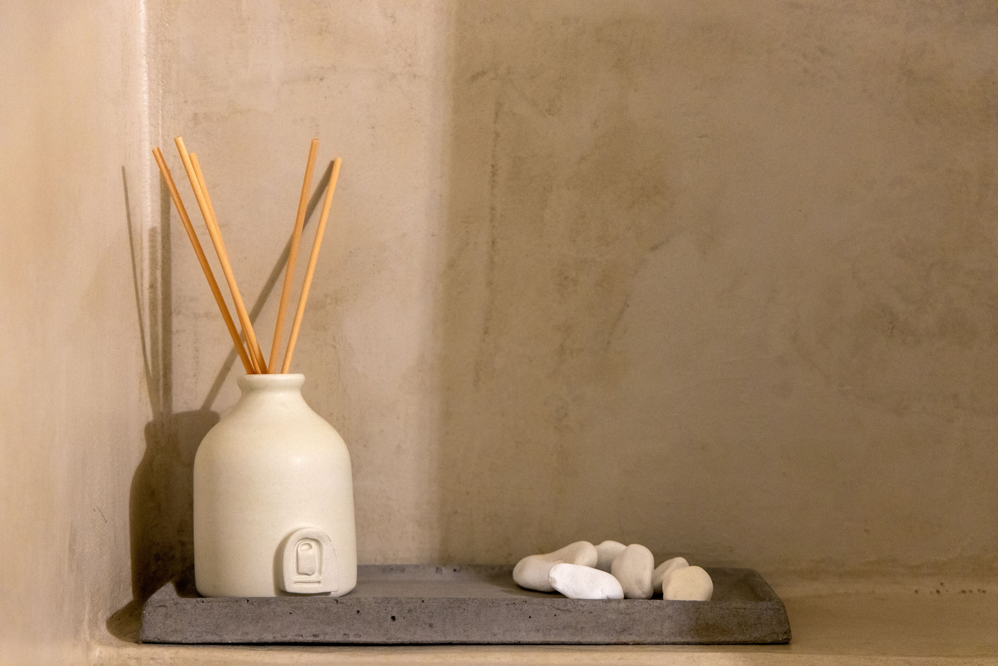 A white ceramic vase with reed sticks on a gray shelf against a textured wall.
