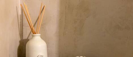 A white ceramic vase with reed sticks on a gray shelf against a textured wall.