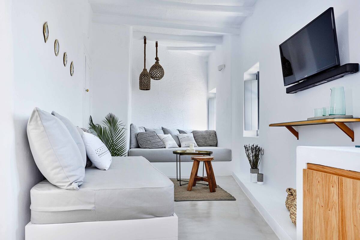 Een moderne woonkamer met een grijze bank, een klein houten krukje, een aan de muur bevestigde televisie en een plant op de bijzettafel.
