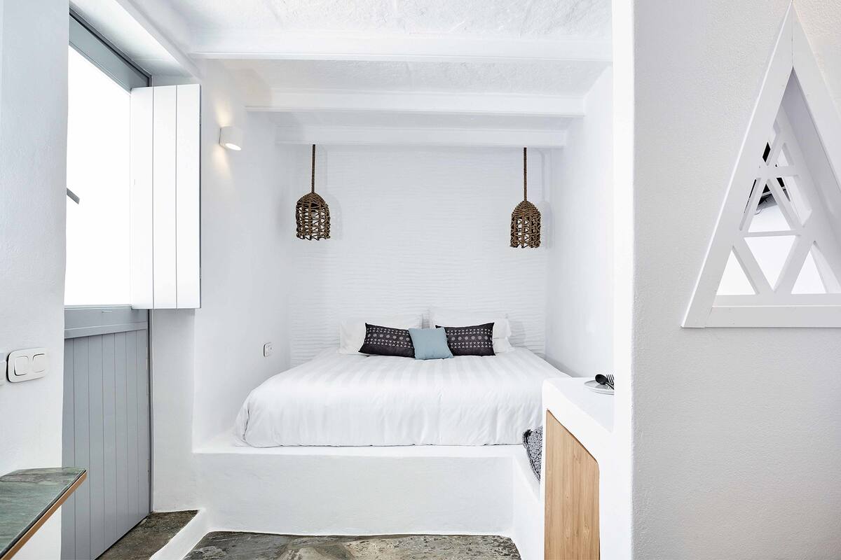 Een slaapkamer met een wit bed, twee hanglampen en een raam met een witte omlijsting.