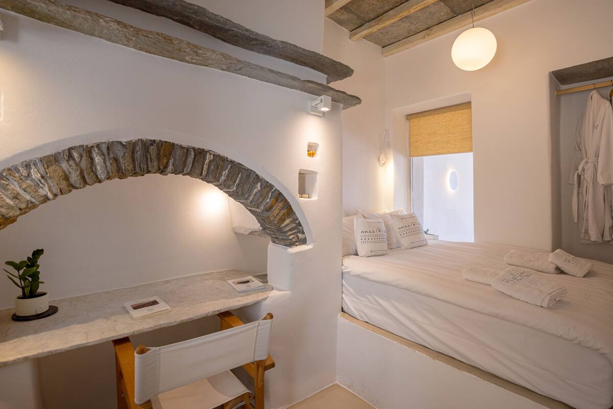Een slaapkamer met een stenen boog, een bed met witte lakens, een nachtkastje met een plant en een hangende badjas.