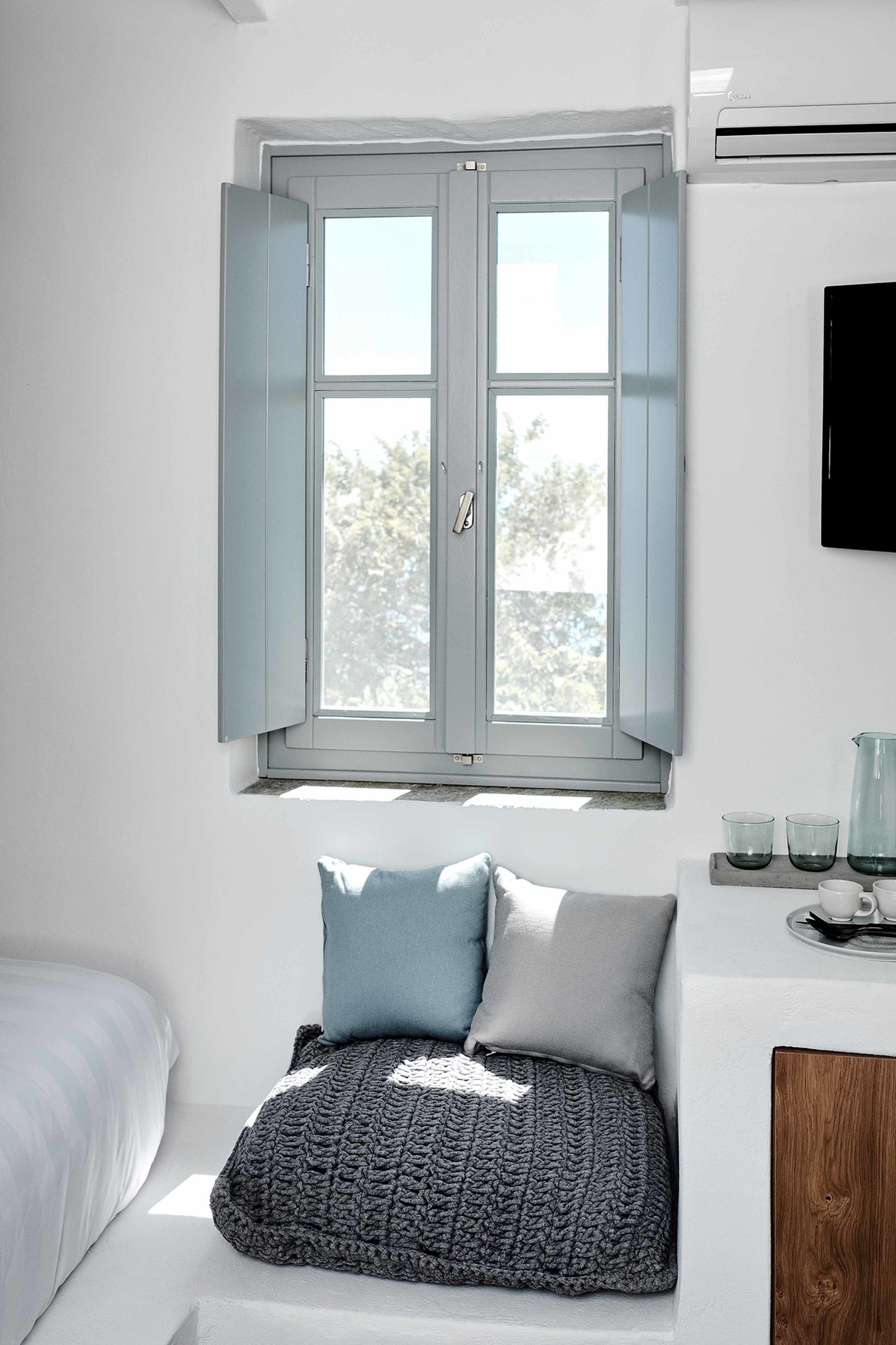 Une chambre à coucher avec un lit, une fenêtre dont les volets sont ouverts, une table de chevet avec une carafe, et un téléviseur fixé au mur.