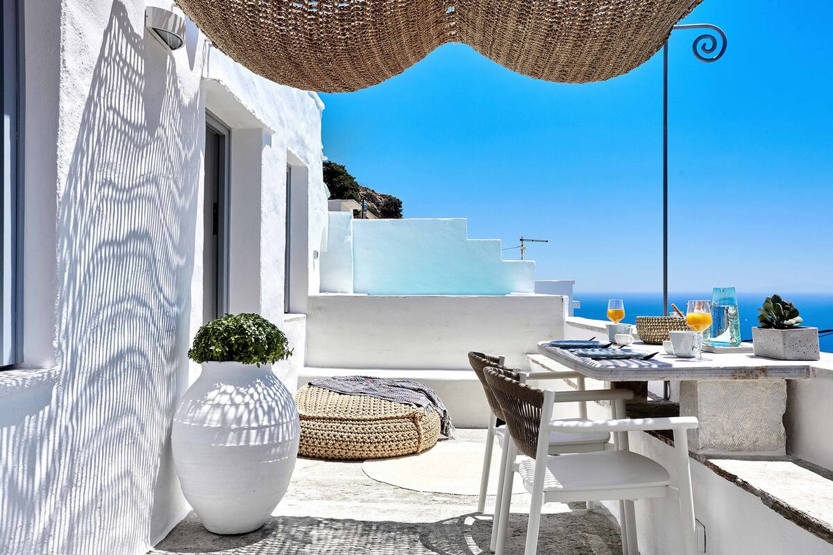 Een terras met witte wanden, een tafel gedekt voor twee personen, een grote potplant en uitzicht op zee.