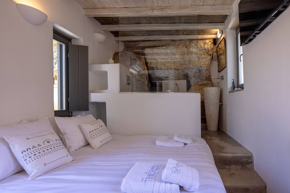 Een slaapkamer met een groot bed, een stenen muur, een kleine badkamer en een raam.