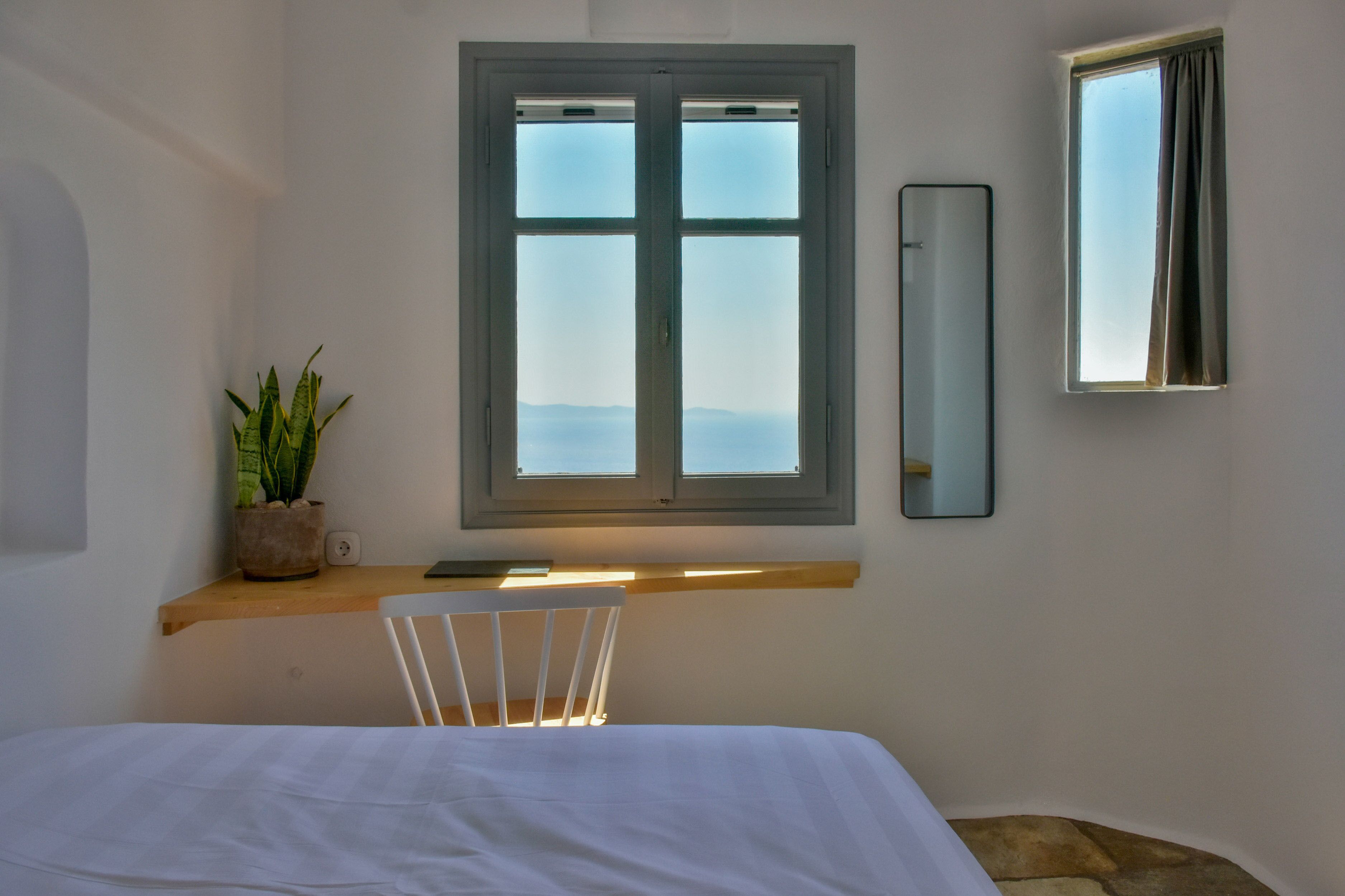 A bedroom with a bed, a window with a view, a bedside table with a plant, and a mirror on the wall.