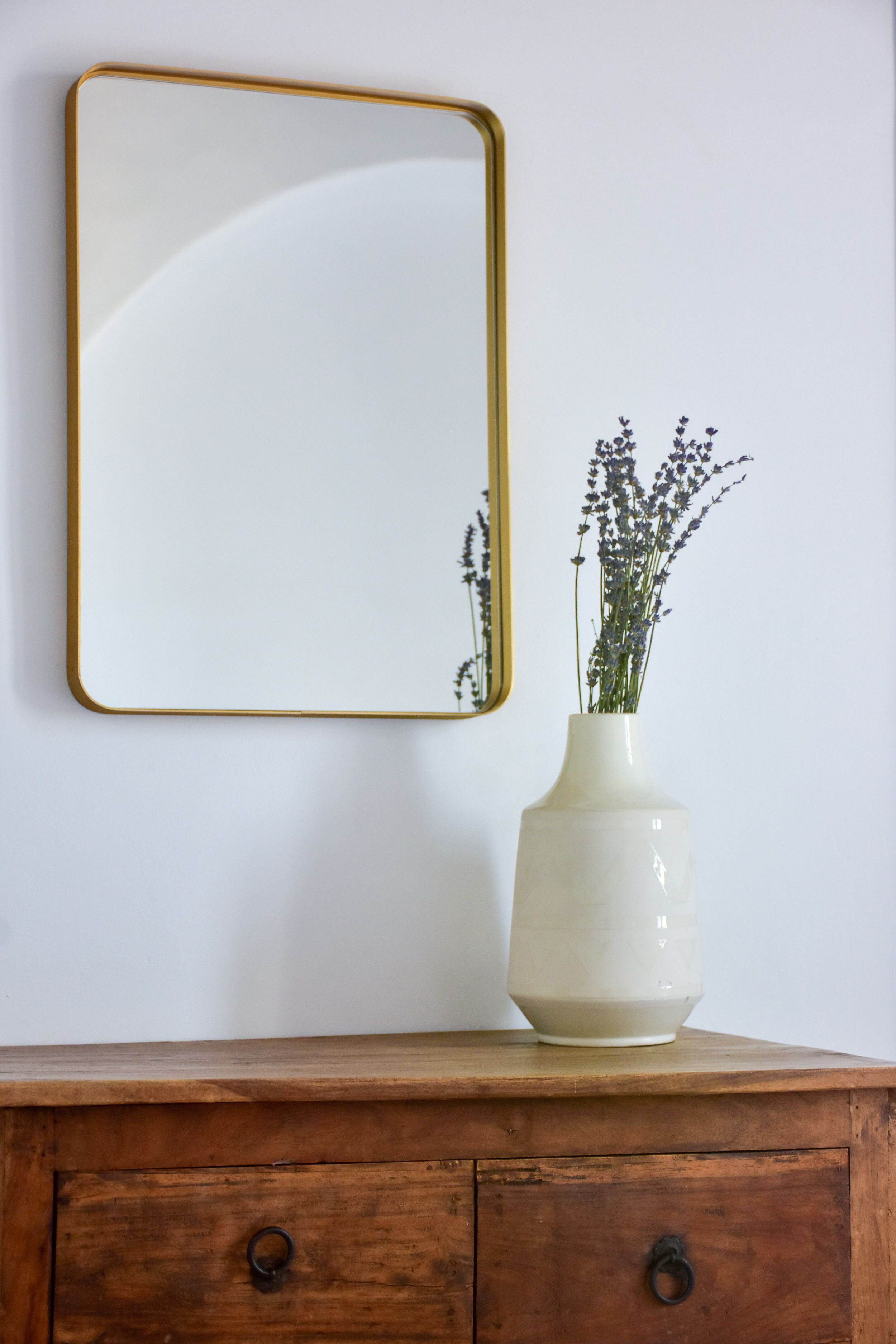 Un jarrón blanco con flores de lavanda sobre un aparador de madera frente a una pared blanca con un espejo con marco dorado.