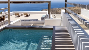 En pool med kristallklart blått vatten, en loungehörna med två solstolar och utsikt över havet och den steniga terrängen.