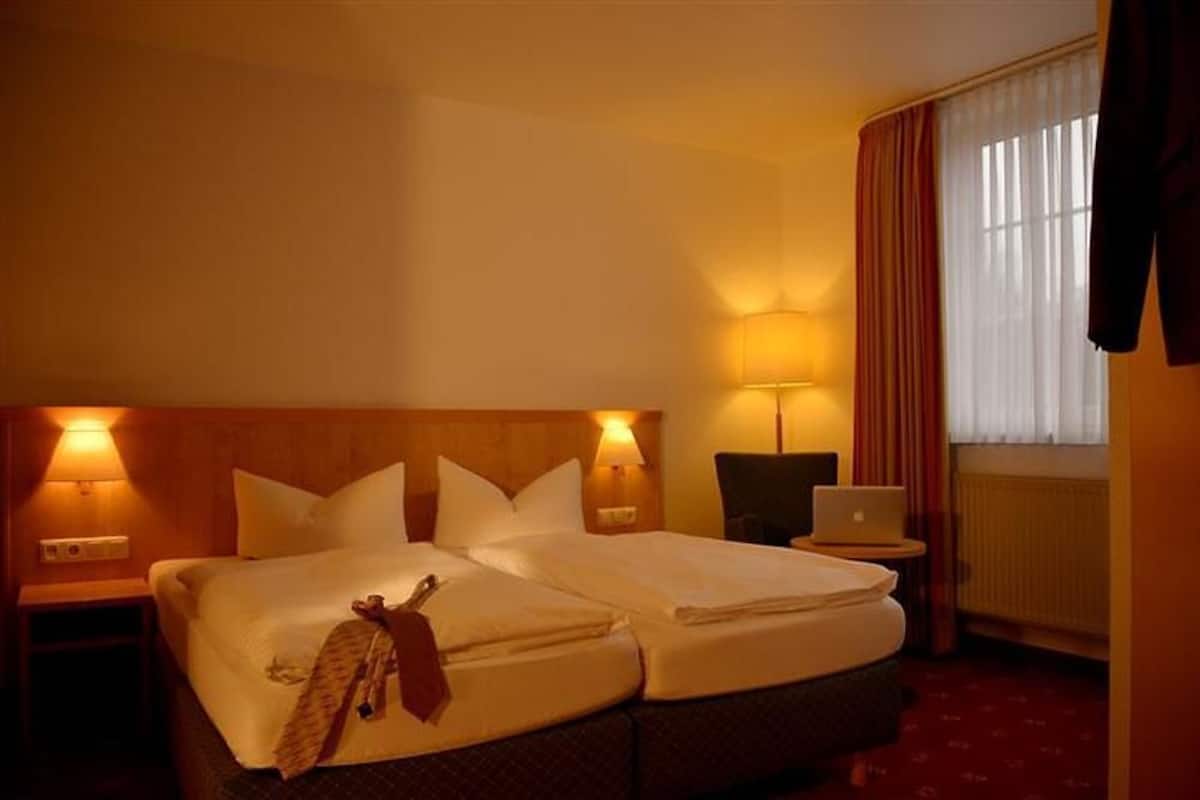 A double bed with a blanket and a tie, two bedside lamps, a laptop on a small table, and a window with curtains.