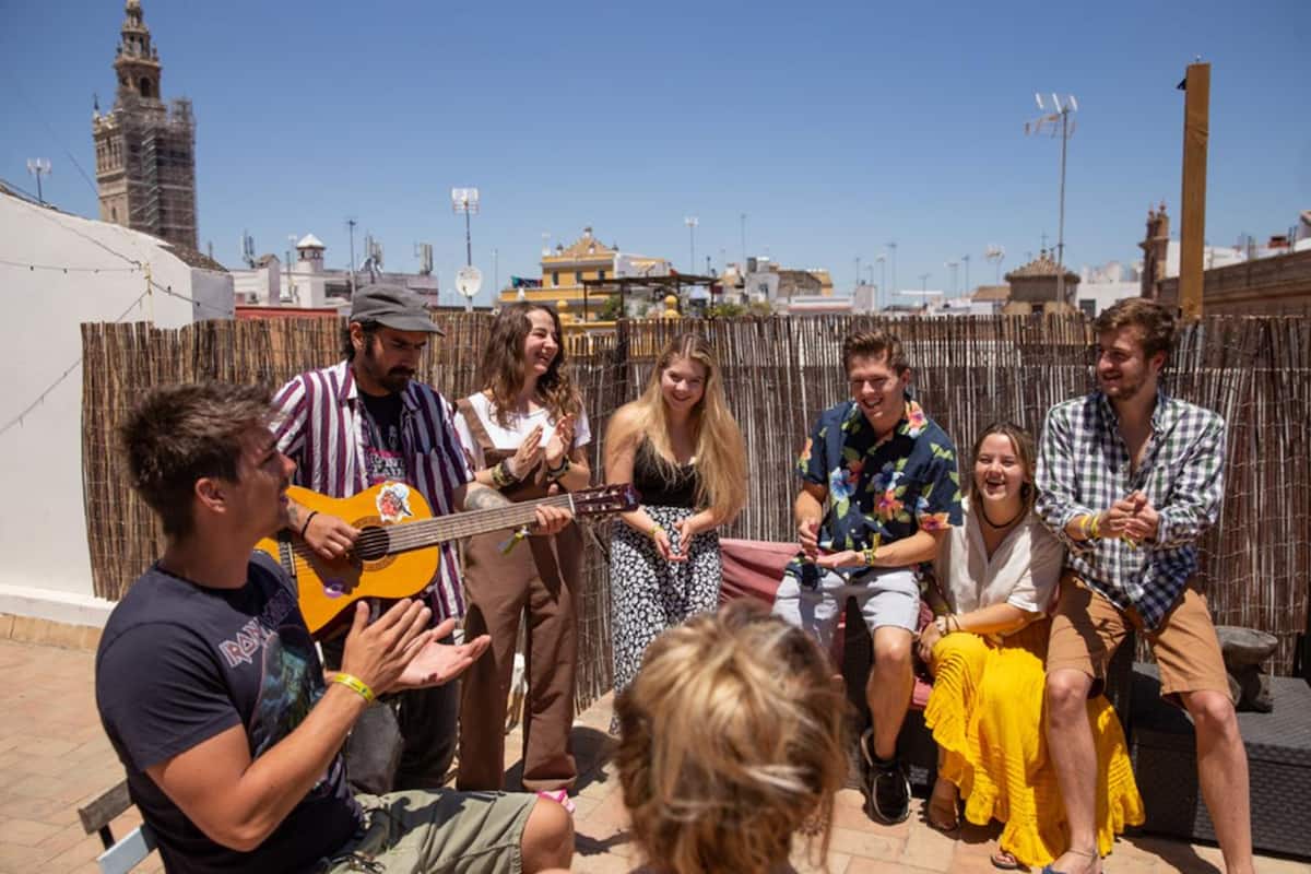 A group of people sitting and standing outdoors, with a musician playing a guitar.