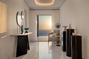 A modern hotel room with a view of the ocean, a sitting area with a chair and a small table, and a large mirror on the wall. - Mykonos Riviera Hotel & Spa a member of Small Luxury Hotels of the World (Mykonos)