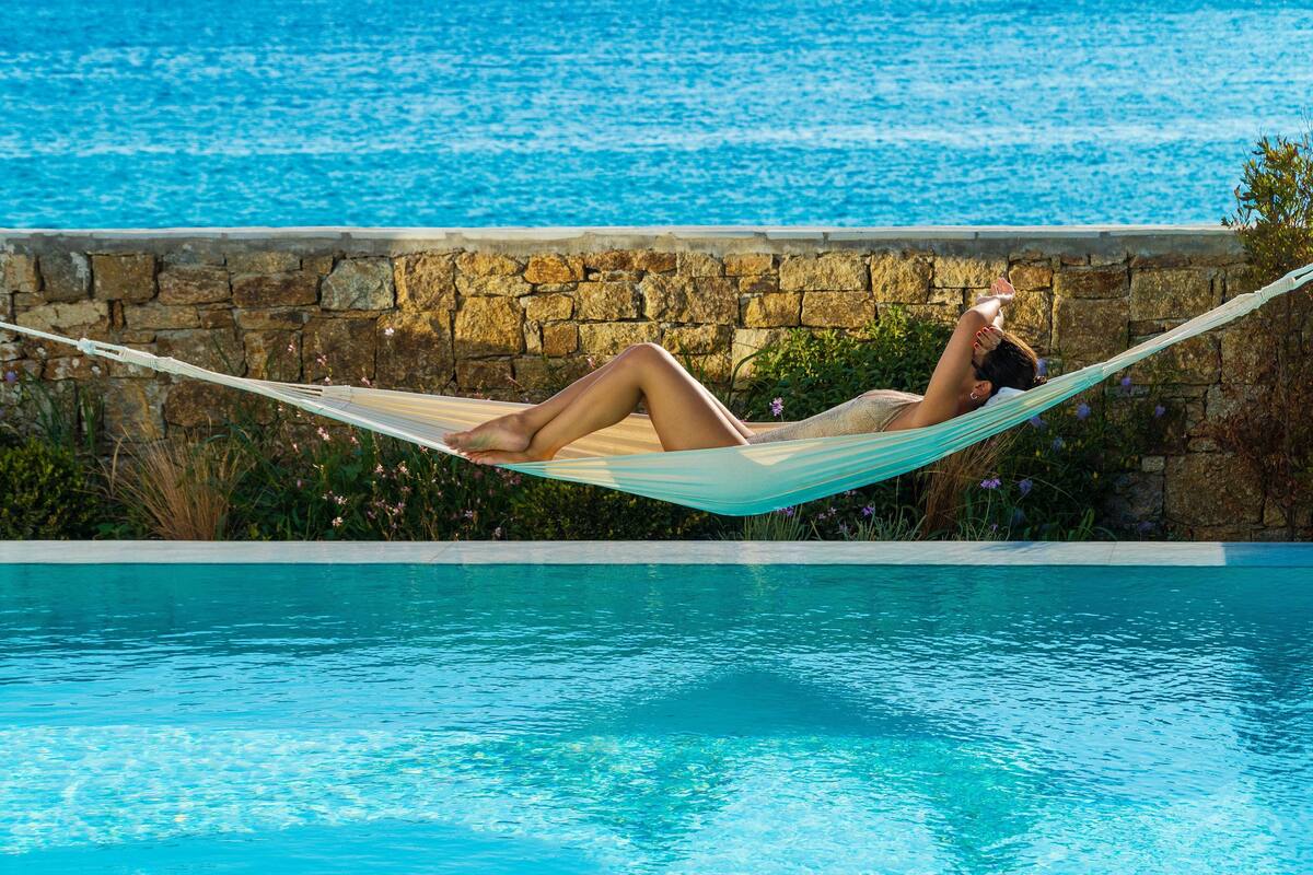A person lying in a hammock by a pool with a stone wall and greenery in the background.