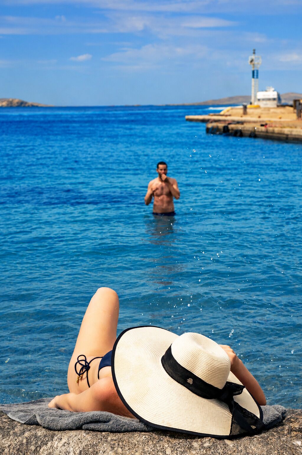 A person relaxing on a beach with a hat, a rocky shoreline, and a lighthouse in the background.