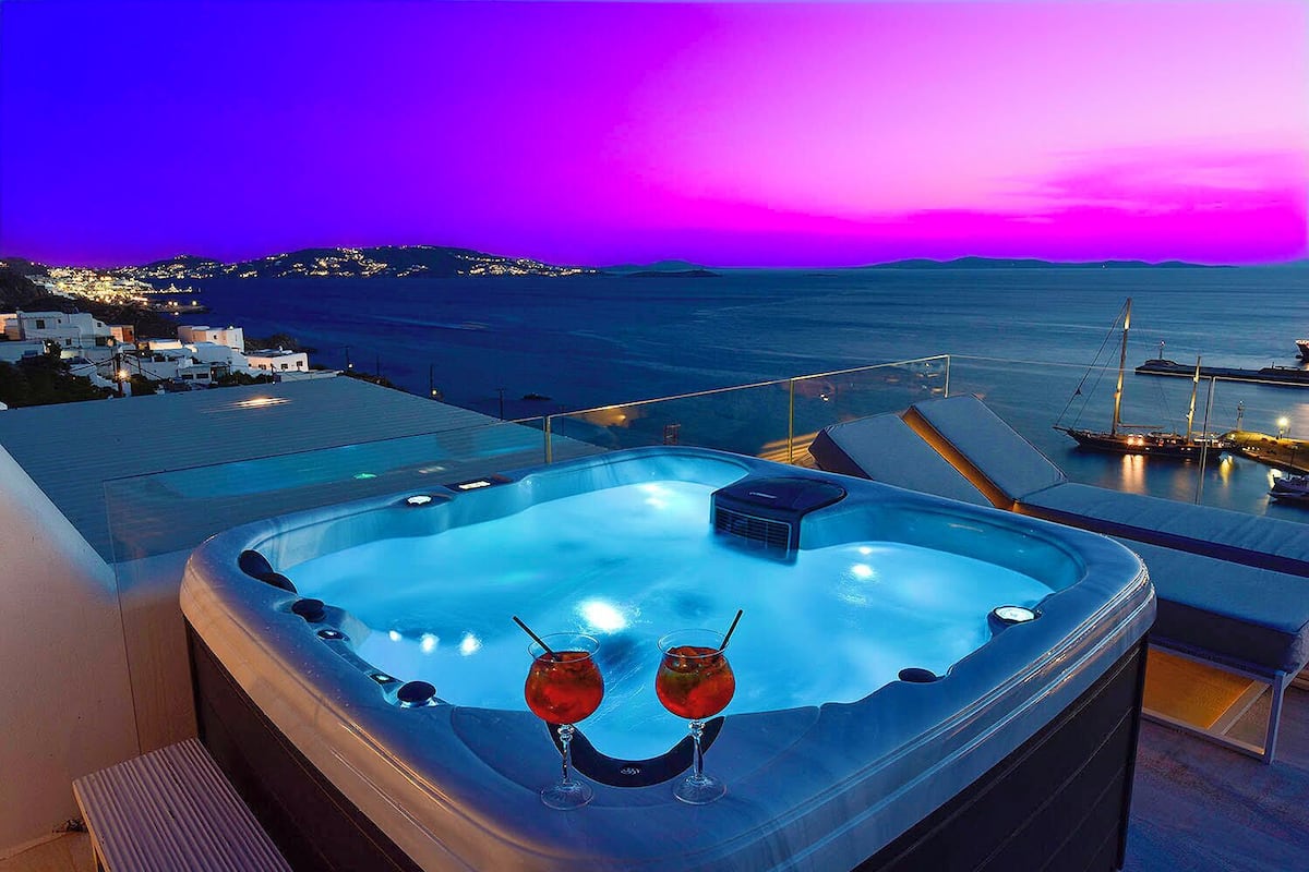 A hot tub with two glasses of red wine, overlooking a marina and a sunset.