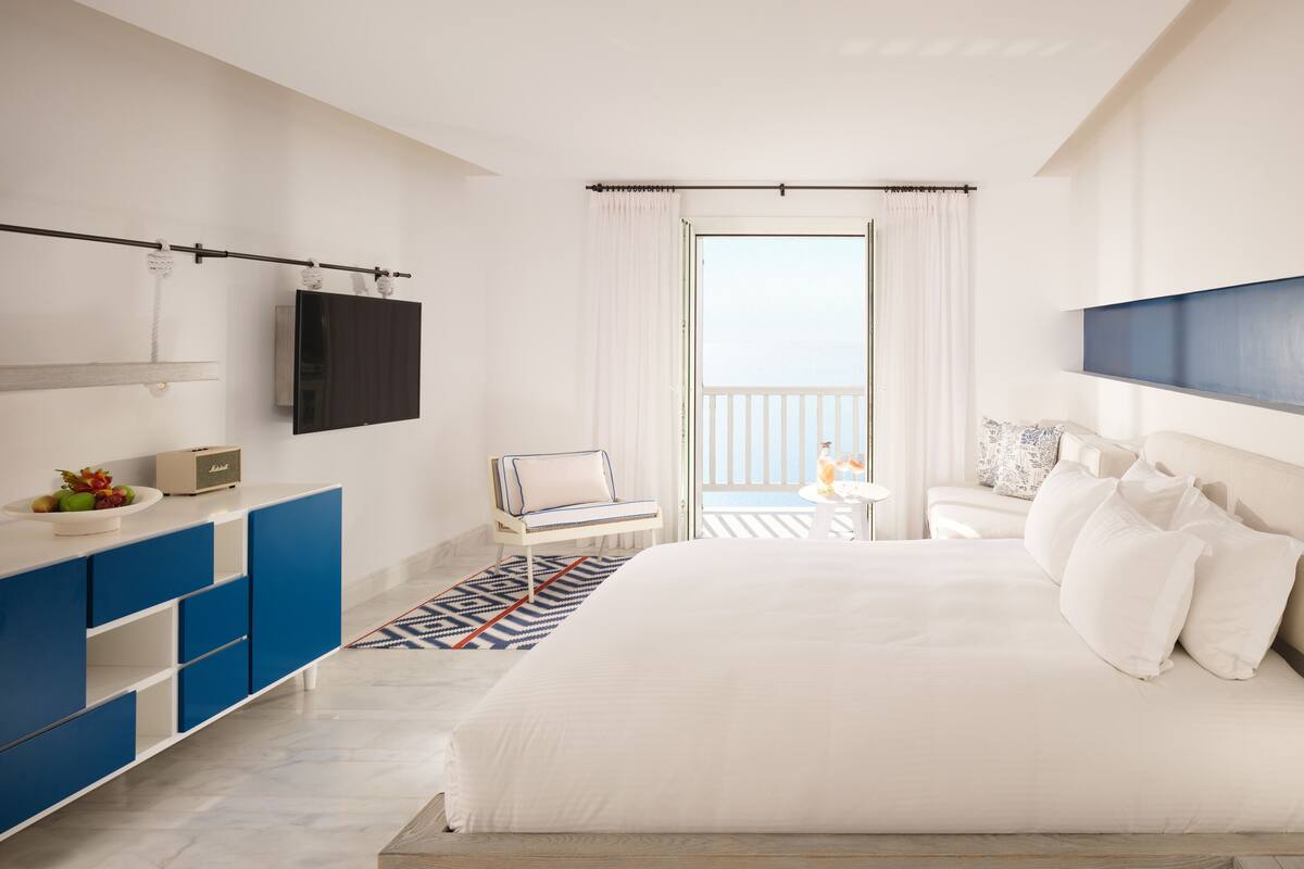 A modern hotel room with a large bed, a TV mounted on the wall, a balcony with a view, and a blue and white patterned rug.