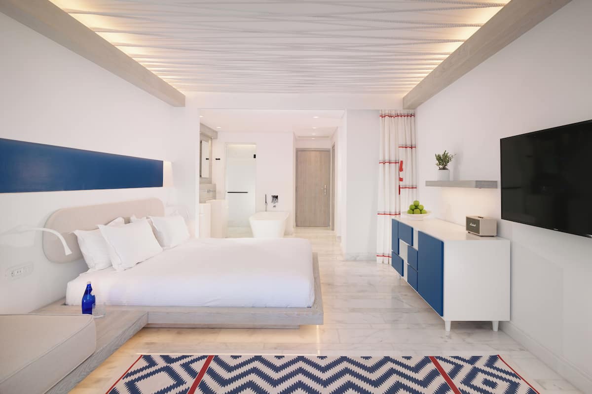 A modern hotel room with a bed, a desk, a TV, and a bathroom visible in the background.