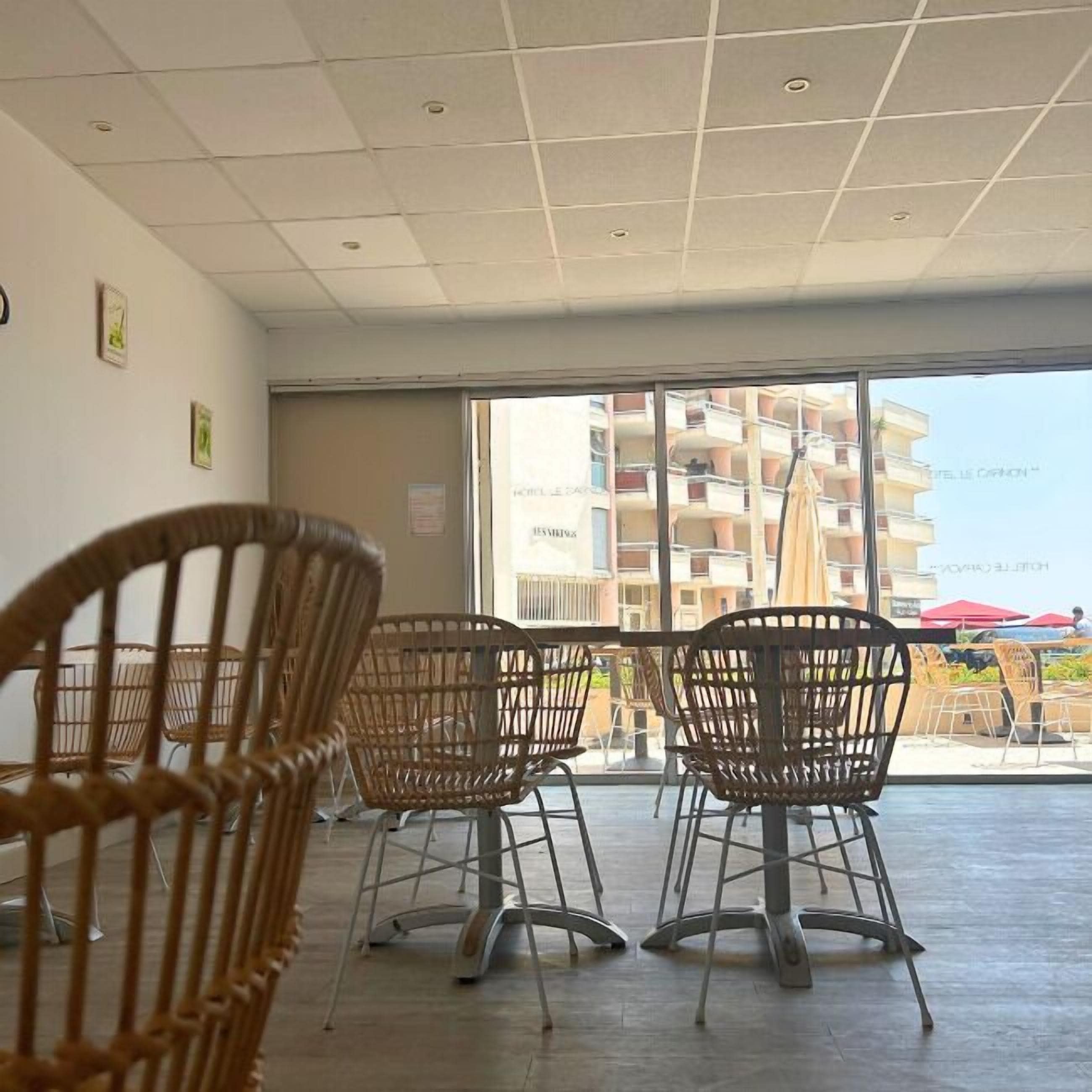 A seating area with wicker chairs and a table, large windows with a view of a building and balconies, and a tiled ceiling with recessed lighting.