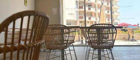 A seating area with wicker chairs and a table, large windows with a view of a building and balconies, and a tiled ceiling with recessed lighting.